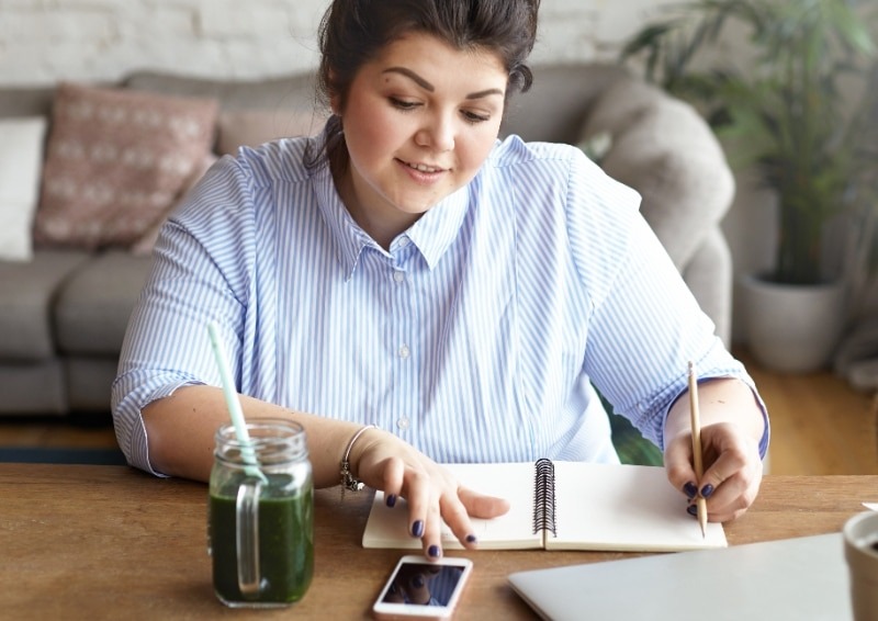 Una mujer sentada en un escritorio, mirando su teléfono inteligente mientras escribe en una libreta.